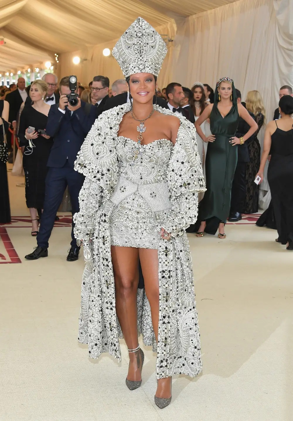 Tại Met Gala 2018 với chủ đề “Heavenly Bodies: Fashion and the Catholic Imagination”, Rihanna đã diện một bộ trang phục, với tưởng được tìm kiếm tại thành phố Rome cổ kính với rất nhiều những di sản nghệ thuật liên quan đến Công giáo ở khắp mọi nơi. NTK John Galliano được tín nhiệm vì những mẫu thiết kế đặc sắc được lấy ý tưởng từ Công giáo mà trước đây ông từng thiết kế khi còn là giám đốc sáng tạo của Dior.
Trang phục được thiết kế bởi John Galliano nhưng Stephen Jones là nghệ nhân đã tạo nên chiếc nón lấy cảm hứng từ vương miện của giáo hoàng Vatican. Túi xách là thuộc thương hiệu Judith Leiber được thiết kế riêng để đồng bộ với trang phục, giày từ Christian Louboutin dòng Pigalle, trang sức vẫn đến từ thương hiệu Cartier. Nữ ca sĩ quả thực đã không khiến nhà tổ chức và người hâm mộ thất vọng khi là khách mời danh dự của sự kiện với bộ cánh được đầu tư ý tưởng và công sức.