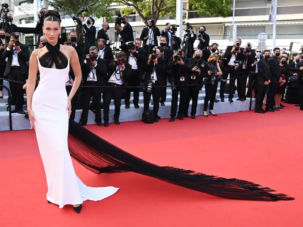 Chiếc đầm Bella Hadid là một sản phẩm "remake" từ thiết kế iconic nằm trong bộ sưu tập Haute Couture năm 2002 của Jean Paul Gaultier. Chiếc váy trắng tinh khôi kết hợp chi tiết cổ chéo với chất vải sheer mang đến sự sang trọng, quyến rũ nhưng không quá hở hang cho người đẹp. Để tổng thể outfit trở nên đặc biệt hơn nữa, nàng siêu mẫu 9x phối cùng khuyên tai đính kim cương và chiếc nhẫn 5,45 carat từ Chopard.