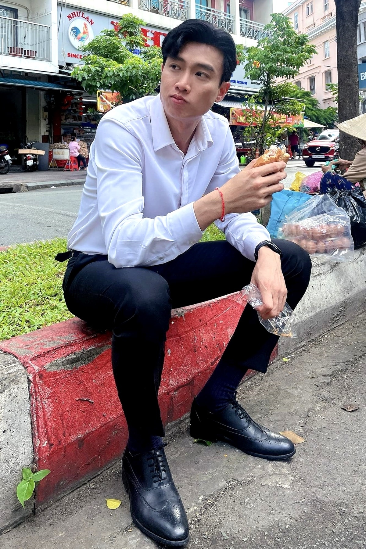 Quốc Trường ngồi bên lề đường ăn bánh mì. “Anh không thích drama, chỉ thích bánh mì và em, vì cả hai đều khiến anh no. Nhưng một cái no bụng, một cái no tình”, nam diễn viên thả thính. (Nguồn: FBNV)