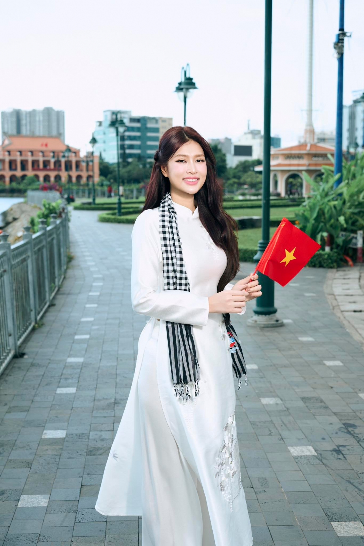 Hoa hậu Đoàn Thiên Ân diện áo dài trắng truyền thống, kết hợp với khăn rằn đậm nét văn hóa Nam Bộ. Trên tay người đẹp cầm lá cờ đỏ sao vàng, nở nụ cười tươi tắn, rạng rỡ.