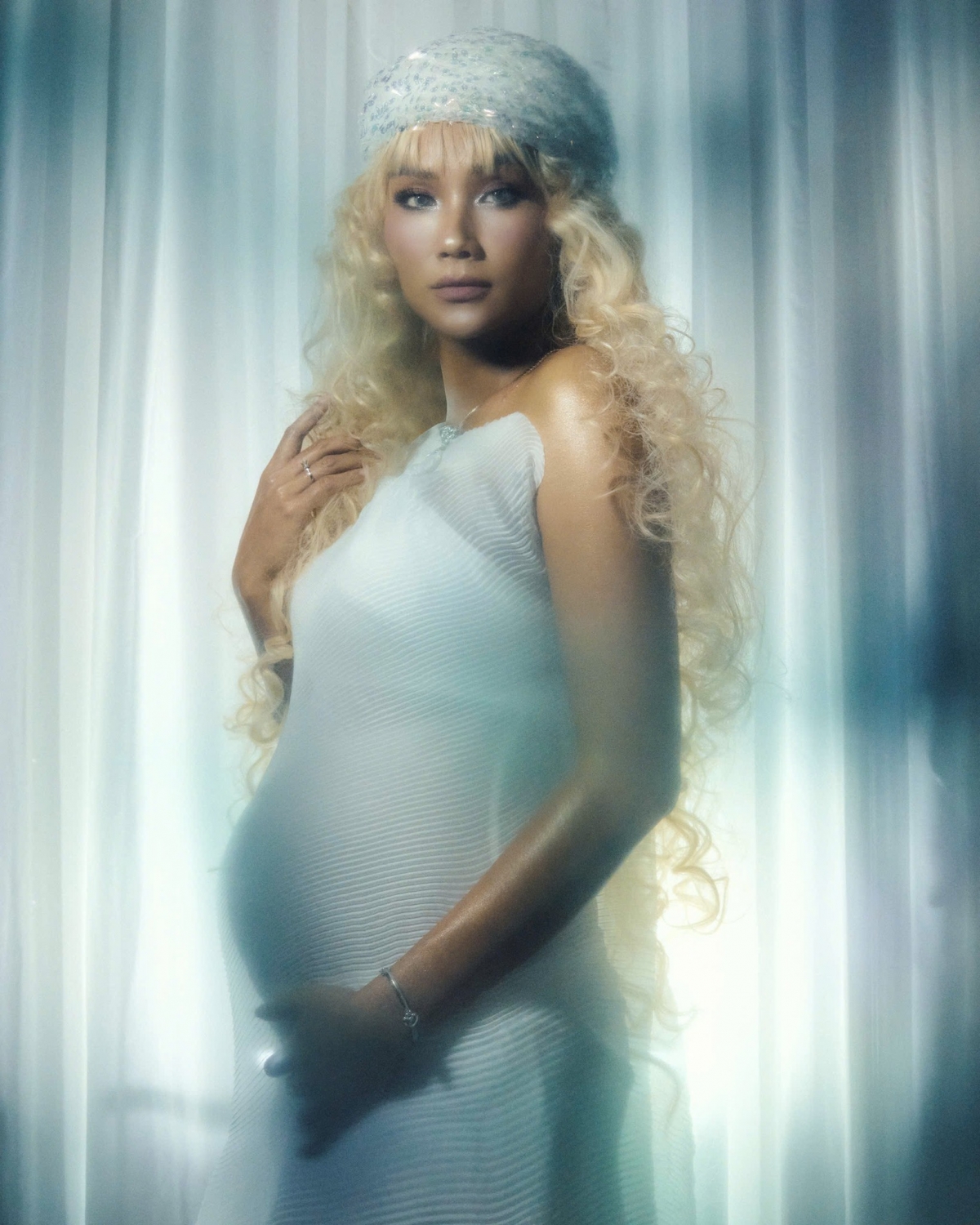 Trong một bộ ảnh, Hoa hậu H'Hen Niê gây ấn tượng khi diện một chiếc váy ôm sát cơ thể, chất liệu mỏng, giúp tôn lên đường cong của cơ thể và khoe trọn bụng bầu. Điểm nhấn của bộ trang phục là mái tóc dài xoăn màu vàng, kết hợp cùng mũ đội đầu đính sequin lấp lánh.