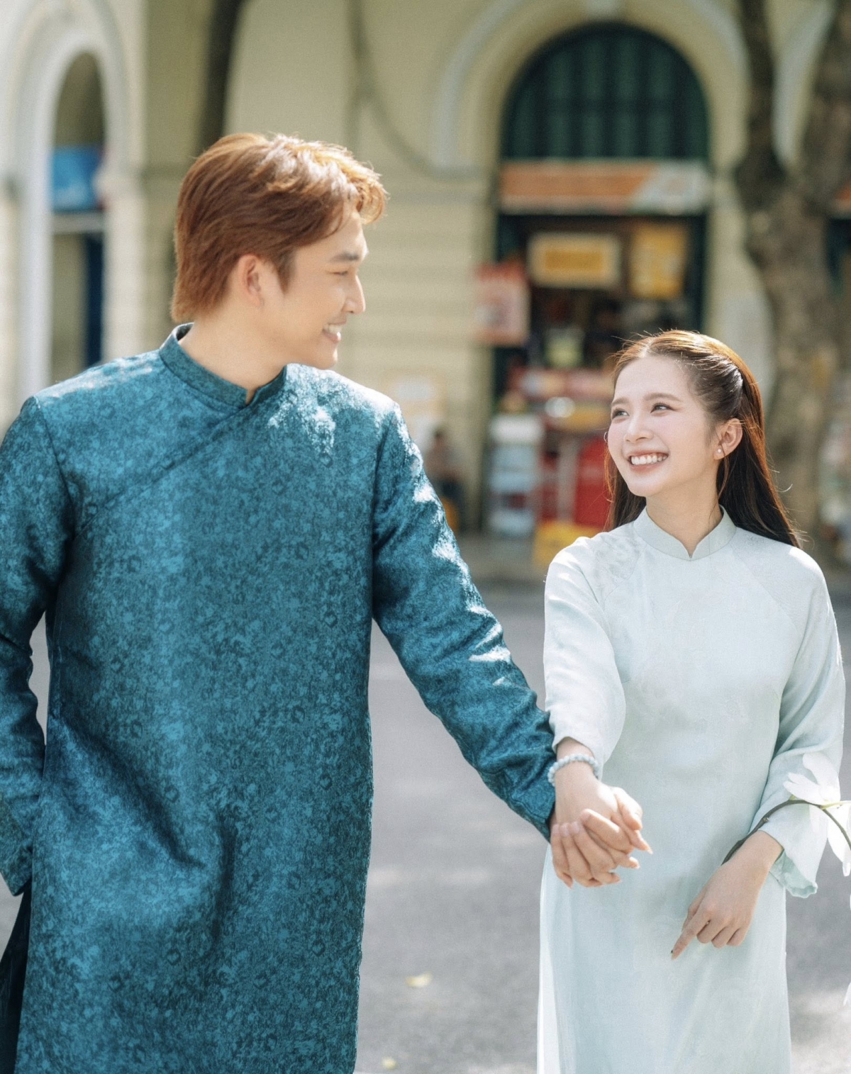 Cả hai diện áo dài gấm truyền thống tông xanh pastel. Chú rể chọn áo dài xanh ngọc đậm kết hợp quần âu đen; cô dâu diện áo dài xanh bạc nhẹ với họa tiết gấm tinh tế, tay cầm bó hoa trắng tối giản.