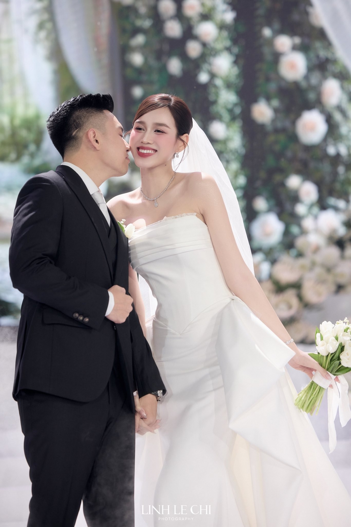 Đỗ Thị Hà diện chiếc váy cúp ngực, xòe bồng để đón khách dự tiệc. Chiếc váy giúp người đẹp khoe vai trần mảnh mai. Chú rể Nguyễn Viết Vương diện suit bảnh bao. Anh nắm chặt tay vợ, cùng cô đón quan khách.