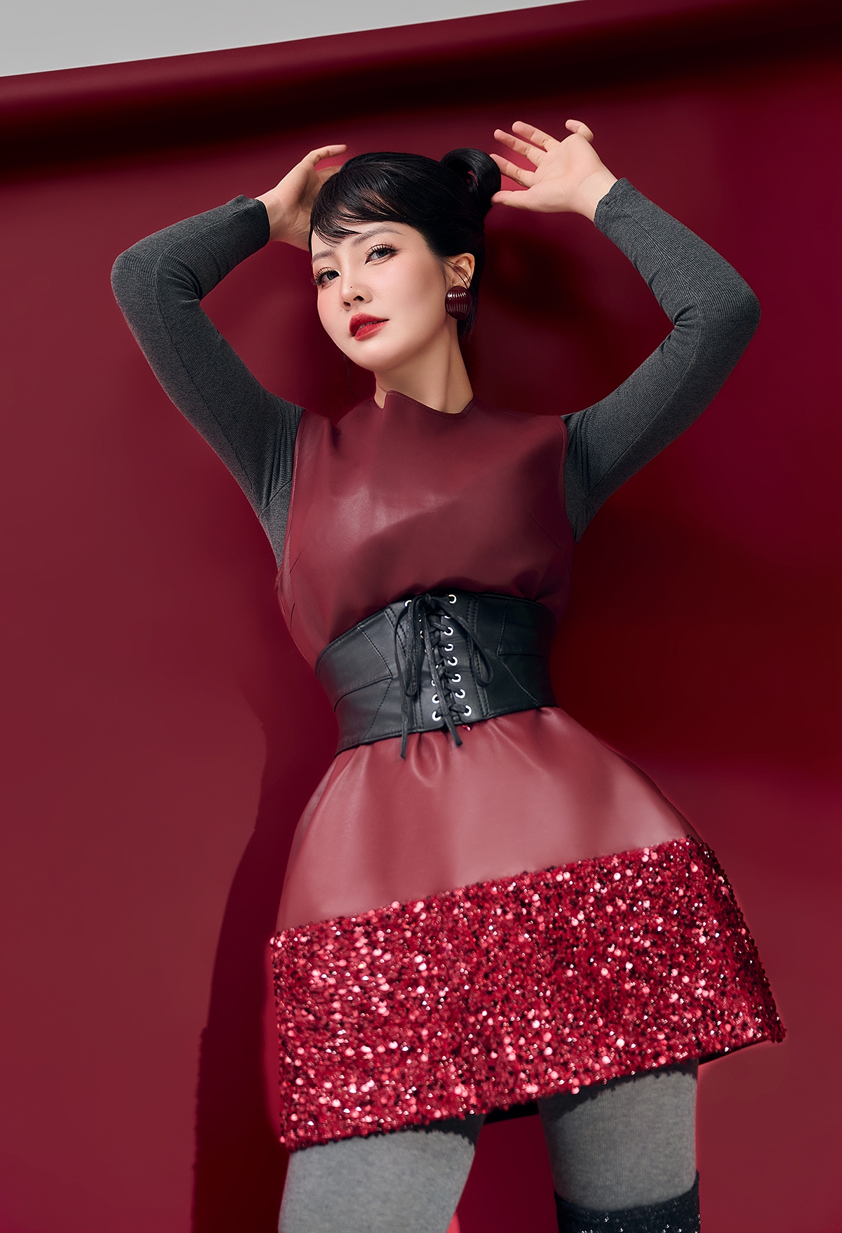 Sắc đỏ đậm bao trùm khung hình không chỉ tạo hiệu ứng thị giác mạnh, mà còn gợi liên tưởng đến sự sống, bản năng và nguồn năng lượng nữ tính của người phụ nữ sau sinh. Trang phục trong bộ ảnh nền đỏ được xử lý theo lối layer tinh tế: sắc đỏ burgundy của chất liệu da mềm và sequin kết hợp cùng tông xám trung tính, đi kèm boots đen lấp lánh.