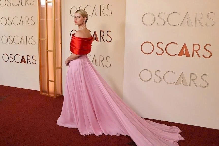 Jessie Buckley mặc chiếc váy hở vai màu đỏ - hồng, với phần tà mềm mại đem lại cảm giác nữ tính, quyến rũ.