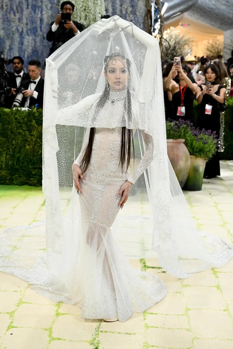 Met Gala 2026 diễn ra tối 4/5 tại New York, Mỹ, tức sáng 5/5 theo giờ Hà Nội, tiếp tục là một trong những sự kiện thời trang được chú ý nhất hành tinh. Năm nay, sự kiện có chủ đề “Costume Art”, với dress code “Fashion Is Art”, gắn liền với triển lãm của Viện Trang phục thuộc Bảo tàng Nghệ thuật Metropolitan. Beyoncé đồng chủ trì dạ tiệc cùng Nicole Kidman, Venus Williams và Anna Wintour. Lisa là gương mặt gây chú ý khi xuất hiện trong bộ đầm đuôi cá trắng được thiết kế riêng bởi Robert Wun. Theo Vogue, nữ ca sĩ hợp tác cùng nhà thiết kế để tạo nên mẫu váy dạ hội mỏng, mang tinh thần kết hợp giữa thời trang và nghệ thuật, đồng thời tôn vẻ đẹp hình thể. Thiết kế lấy cảm hứng trực tiếp từ chủ đề “Costume Art” và chính cơ thể của Lisa. Đội ngũ nhà mốt đã quét 3D cánh tay của cô để mô phỏng các tư thế múa truyền thống Thái Lan, tạo nên chi tiết hai cánh tay đính ở phần cầu vai và nâng lớp voan phía sau.